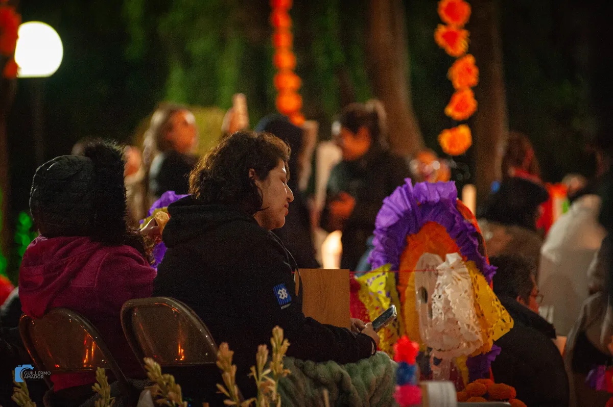 Personas sentadas de noche entre adornos de papel de colores y guirnaldas de flores naranjas, durante una celebración de Día de Muertos.