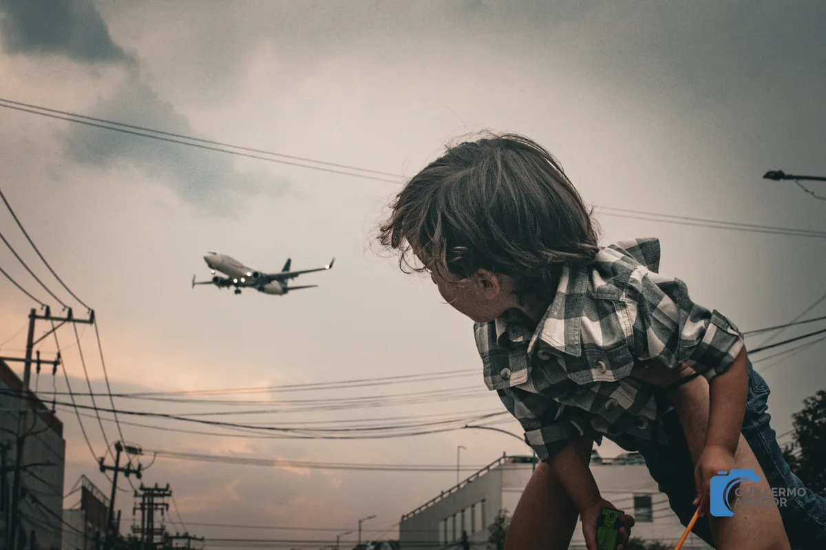 Niño con camisa a cuadros mirando un avión que pasa en el cielo nublado, entre cables eléctricos y edificios.