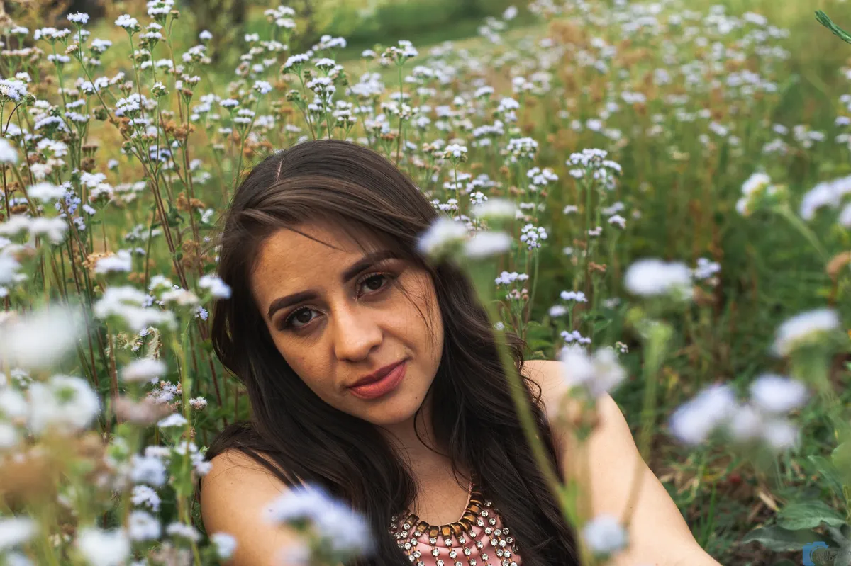 Mujer de cabello largo oscuro mirando a la cámara entre flores silvestres blancas.