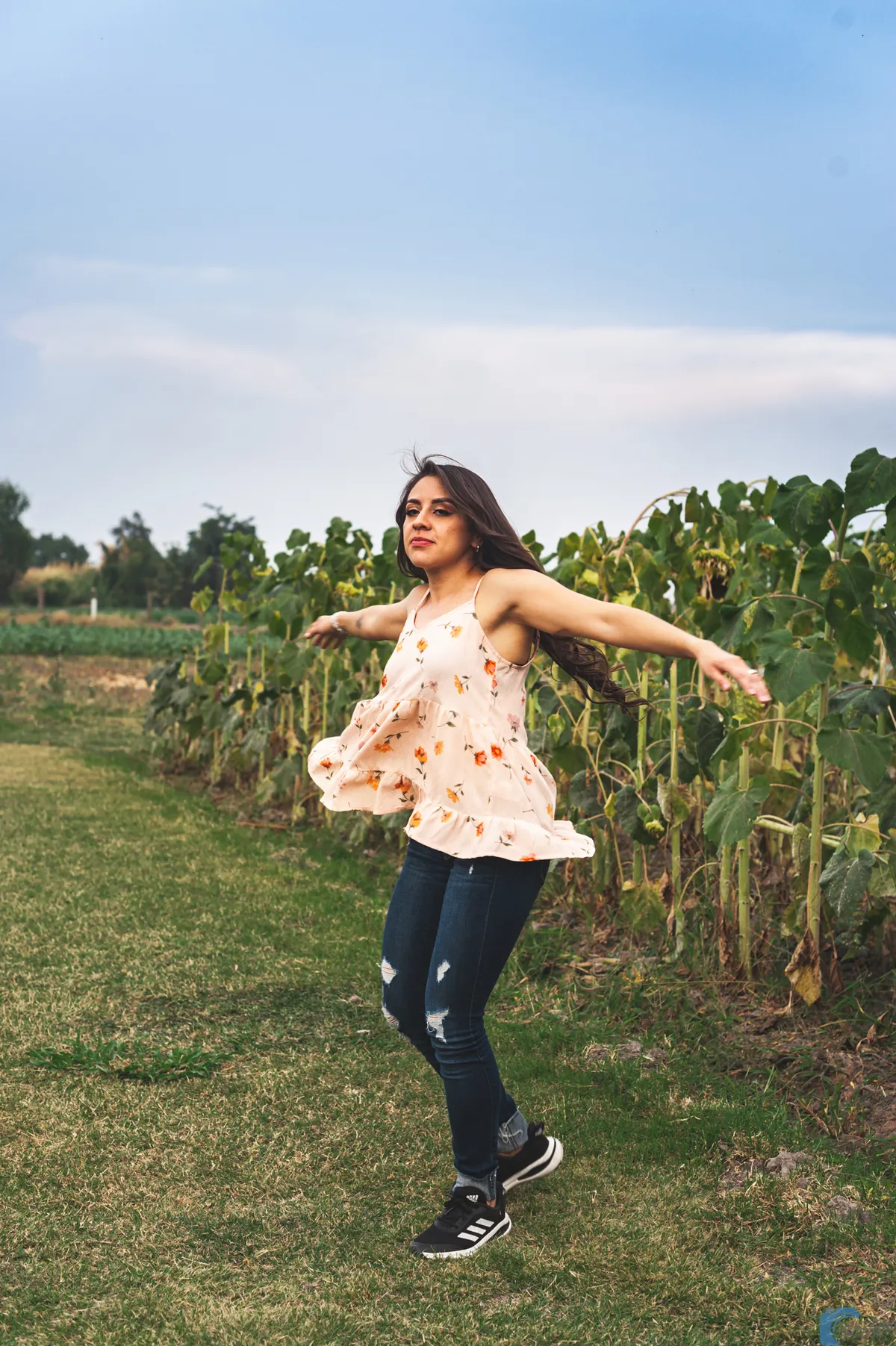 Mujer con blusa clara estampada y jeans girando con los brazos abiertos sobre el pasto, junto a un campo de cultivo.