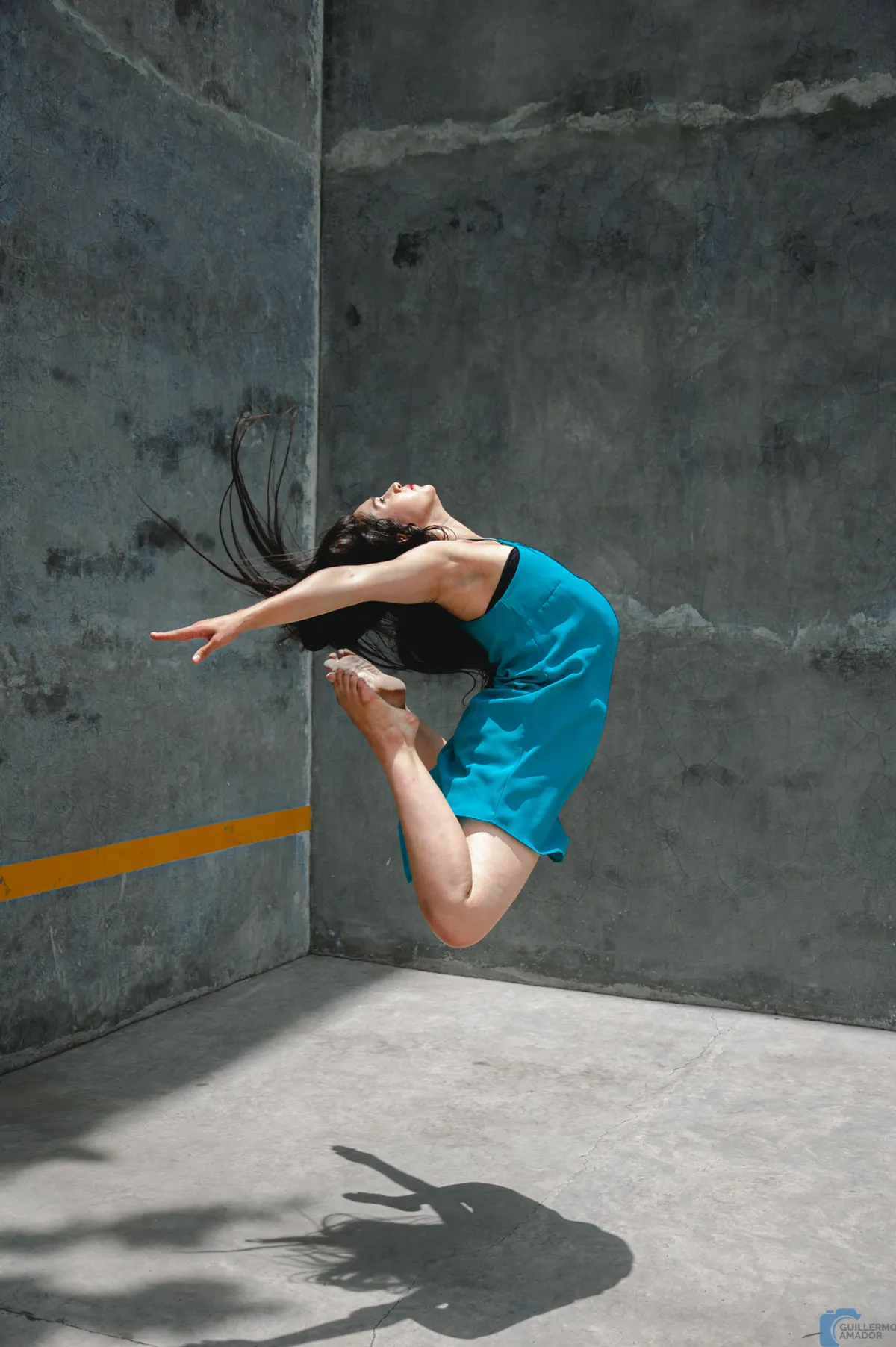Mujer con vestido turquesa en pleno salto, con la espalda arqueada, frente a muros de concreto; su sombra se proyecta en el piso.
